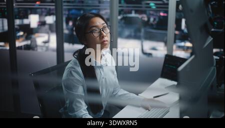 Asian Female Financial Specialist Sits at Innovative Workspace, Eyes Scanning Multiple Displays Filled With Trading Indices. Young Lady Actively Uses Keyboard to Input Data and Execute Market Orders. Stock Photo