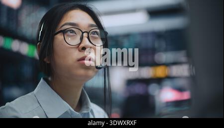 Close Up of Young Asian Woman in Headset Analyzing Charts, Executing Strategic Buy and Sell Orders, Managing Stocks and Digital Assets With Precision, Confidence in Fast Paced Financial Environment. Stock Photo