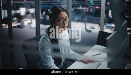 Asian Female Financial Specialist Sits at Innovative Workspace, Eyes Scanning Multiple Displays Filled With Trading Indices. Young Lady Actively Uses Keyboard to Input Data and Execute Market Orders. Stock Photo