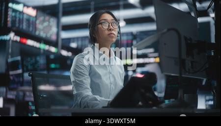 Female Trader Analyzing Real Time Stock Market Data at Large Screens. Focused Asian Woman in Glasses Works Intently at High Tech Desk, Typing on Keyboard While Monitoring Complex Financial Charts. Stock Photo