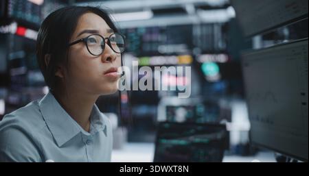 Professional Asian Female Trader Works in High Tech Digital Trading Center, Carefully Monitoring Live Market Fluctuations Across Screens. Wearing Headset, Girl Executes Strategic Buy and Sell Orders. Stock Photo
