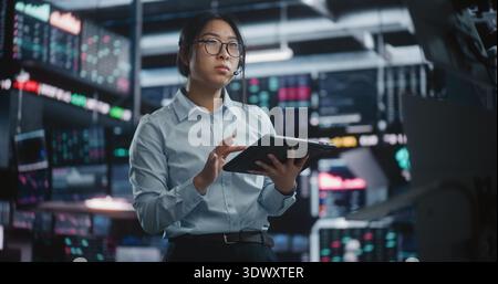 Asian Financial Specialist Consulting Client Via Headset While Syncing Tablet and Screen Analytics. Strategic Thinking and Integration of Mobile and Desktop Analytics in Futuristic Stock Exchange Hub. Stock Photo
