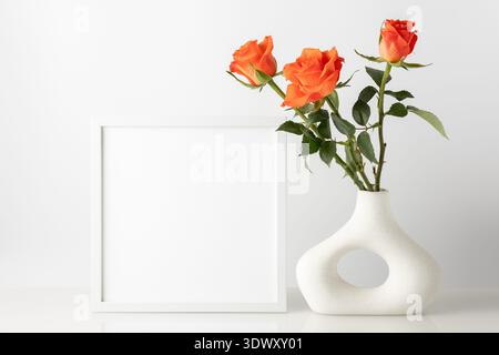 Bouquet of natural coral orange rose flowers in modern donut shape vase and empty photo frame on glass table against white wall. Home interior decorat Stock Photo
