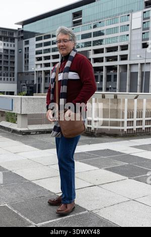 Senior man standing on paved terrace by modern building, wearing red sweater, carrying brown bag Stock Photo