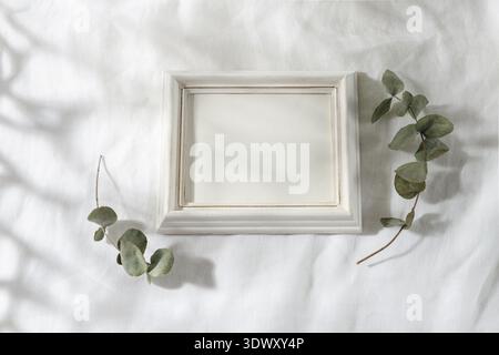 Empty horizontal retro white wooden photo or picture frame with  dried eucalyptus branches on linen tablecloth in sunlight. Summer minimal still life Stock Photo