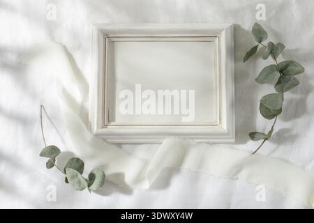 Empty horizontal retro white wooden photo or picture frame with silk ribbon and dried eucalyptus branches on linen tablecloth in sunlight. Summer mini Stock Photo
