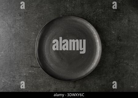 Empty grunge dark grey craftmade ceramic plate on black stone board background. Retro style dishware. Stock Photo