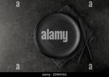 Empty grunge dark grey craftmade ceramic plate with cutlery and linen serviette on black stone board background. Retro style table serving matte dishw Stock Photo