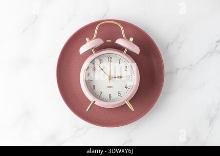Pink alarm clock on retro red plate on white marble table background. Time to food. Meal on time as element of healthy diet. Stock Photo