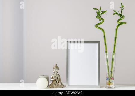 Floral card or poster mockup or silver photo frame with bamboo plant in glass vase,  decorative Buddha statuette and candles on white tablecloth. Home Stock Photo