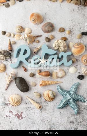 Wooden blue word Relax, ceramic starfish and various colorful natural seashells and pebbles scattered on grunge stone background. Summer vacation conc Stock Photo