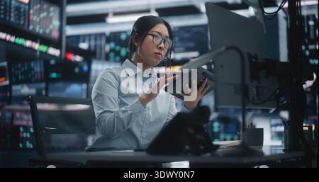 Focused Asian Female Trader Analyzing Market Trends via Tablet and Multi Monitor Setup. Woman in Glasses Sits at Workstation, Cross Referencing Real Time Data on Digital Tablet and Talking on Headset. Stock Photo