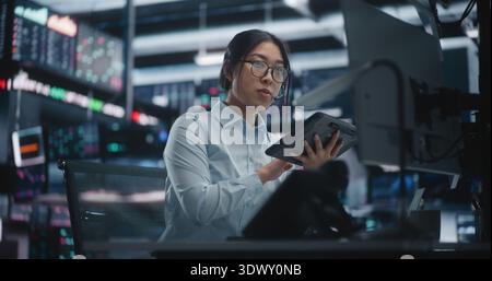 Female Expert Investor Monitoring Real Time Data and Communicating in Futuristic Hub. Young Lady Examines Intricate Stock Charts on Tablet While Discussing of Investment Strategies Through Headset. Stock Photo