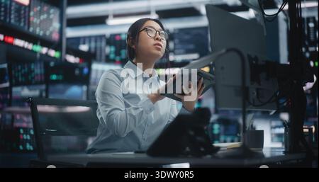 Focused Asian Female Trader Analyzing Market Trends via Tablet and Multi Monitor Setup. Woman in Glasses Sits at Workstation, Cross Referencing Real Time Data on Digital Tablet and Talking on Headset. Stock Photo