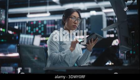 Female Expert Investor Monitoring Real Time Data and Communicating in Futuristic Hub. Young Lady Examines Intricate Stock Charts on Tablet While Discussing of Investment Strategies Through Headset. Stock Photo