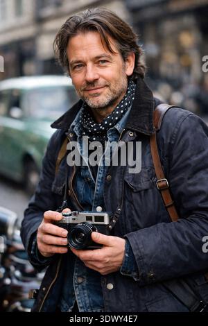 A rugged street photographer stands on a city street, wearing a classic navy waxed cotton jacket  holding a vintage silver and black rangefinder photo Stock Photo