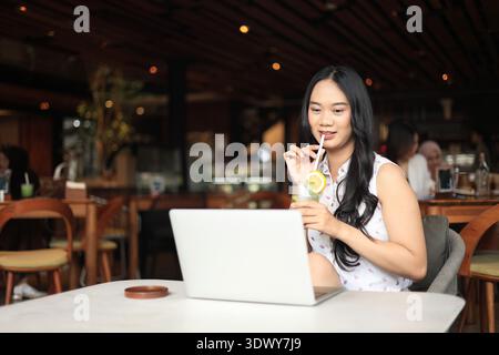 Young Asian Freelancer Enjoying a Healthy Fresh  Juice while Working Remotely on Her Laptop in a Modern Cafe or Coffeeshop. Work from Cafe Concept Stock Photo