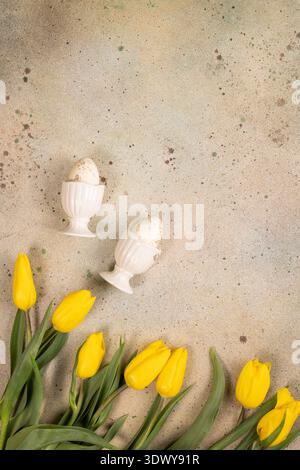 Speckled easter eggs nestled in white eggcups, surrounded by vibrant yellow tulips arranged on a rustic concrete background, creating a festive spring Stock Photo
