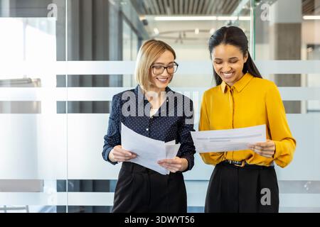 Two smiling diverse businesswomen standing and collaborating in a modern office hallway, discussing project documents and planning cheerfully with a blurred glass wall background Stock Photo