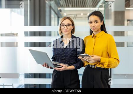 Two confident businesswomen, one holding a laptop and the other a tablet, standing together in a modern glass office, representing teamwork and professional partnership Stock Photo
