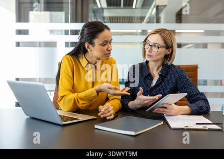 Two diverse businesswomen actively collaborating and communicating during a corporate meeting, one holding a digital tablet and the other gesturing, emphasizing teamwork and professional discussion Stock Photo