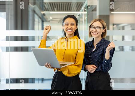 Diverse businesswomen standing in a modern office, one holding a laptop while both show clenched fists, smiling and celebrating a successful project or achievement in their professional work Stock Photo