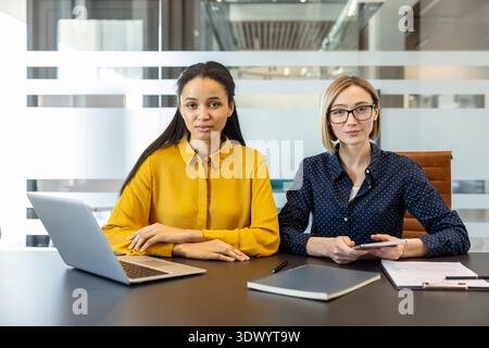 Two diverse businesswomen are sitting at a conference table with a laptop, tablet, and documents, actively engaging in professional collaboration within a modern glass-partitioned office environment Stock Photo