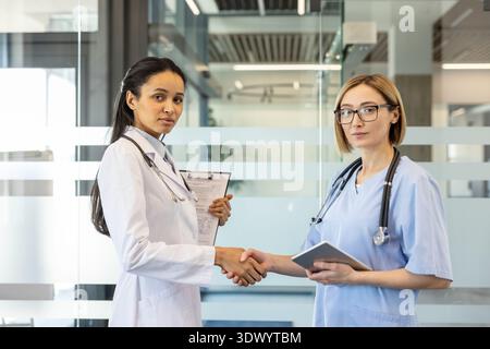 Two diverse female doctors, one holding a clipboard and another a tablet, are shaking hands and signifying agreement, collaboration, and partnership in a modern medical environment Stock Photo