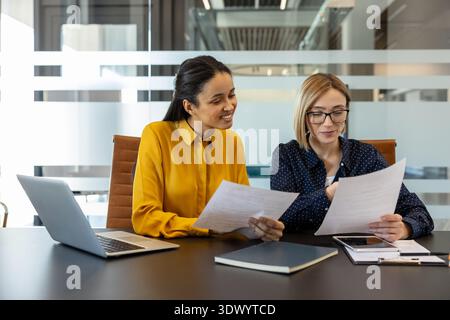 Two professional businesswomen collaborate in a bright modern office, reviewing reports and planning strategy with laptop and tablet, focused, smiling, and productive colleagues Stock Photo