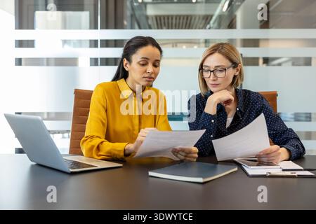 Two diverse businesswomen reviewing documents and discussing work, collaborating on a project or report while sitting at a desk in a modern corporate office environment Stock Photo