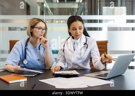 Two diverse female medical professionals are actively engaged in a discussion, reviewing important data on a laptop and documents while working together in a modern hospital office meeting room Stock Photo