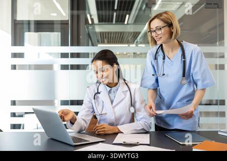 Two smiling multi-ethnic female medical professionals, a doctor and a nurse, discussing patient data and collaborating while looking at a laptop in a hospital office Stock Photo
