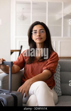 Brazilian woman with glasses sitting on sofa holding suitcase handle Stock Photo