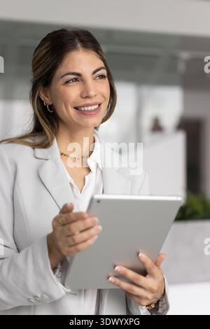 Smiling businesswoman standing in modern office with tablet looking away Stock Photo