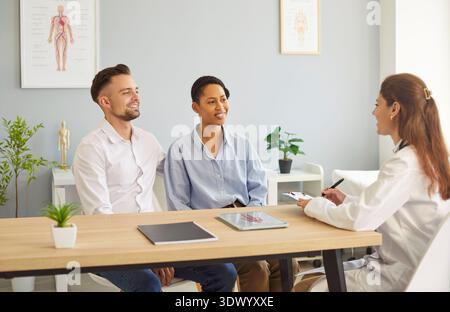 Smiling happy young couple sitting with family doctor therapist in medical clinic. Stock Photo