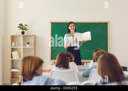 Smiling teacher explaining lesson with open book in classroom to engaged students Stock Photo