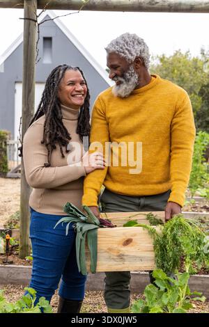 Senior couple wearing sweaters holding wooden crate with leeks carrots in backyard garden Stock Photo