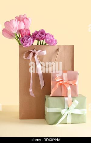 Paper bag with beautiful tulips and gift boxes on yellow background. Mother's Day celebration Stock Photo