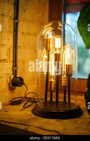 Retro industrial style table lamp with multiple glowing Edison vintage light bulbs under a glass dome on a brick wall windowsill. Stock Photo