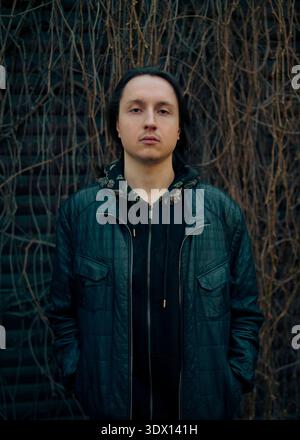 Frontal portrait of a calm, serious caucasian young man with long dark hair wearing a dark blue jacket, standing by a dry vine wall. Stock Photo
