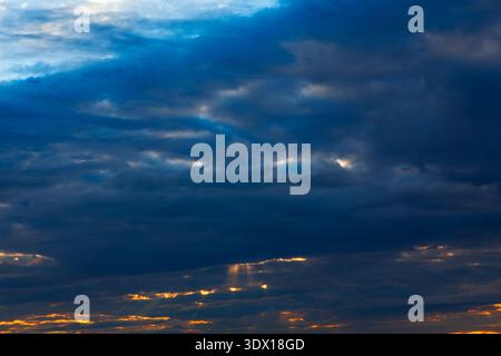 Dramatic sky filled with deep blue and gray clouds captures the fading light of sunset, with golden rays peeking through the breaks. Clouds evoke calm Stock Photo
