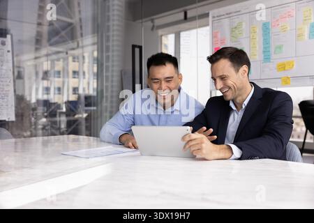 Friendly laughing two diverse businessmen discussing project insights using tablet Stock Photo