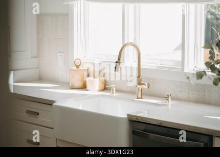 Sunlit kitchen window above farmhouse sink with brass faucet and indoor plants in a bright modern residential kitchen. Stock Photo