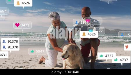 Kneeling couple in polo and tank petting dog at beach, man holding smartphone with social icons Stock Photo