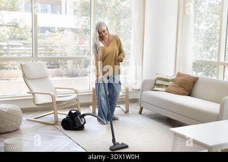 Smiling mature woman with long silver hair vacuuming living room Stock Photo