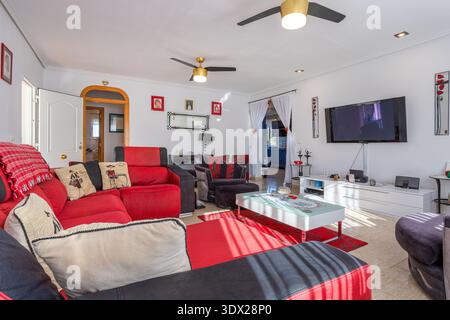 Bright contemporary living room featuring bold red sectional sofa, gray armchairs, glass coffee table, wall mounted television, tiled floor, and elega Stock Photo