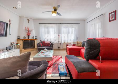 Sunlit modern living room featuring bold red sectional sofa, gray armchairs, fireplace, glass coffee table, tiled floor, and elegant decorative accent Stock Photo