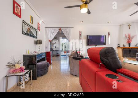 Bright contemporary living room featuring red and gray sectional sofa, wall mounted TV, fireplace, glossy tiled floor, and sheer curtains leading to d Stock Photo