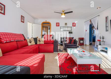 Sunlit contemporary living room featuring bold red sectional sofa, gray armchairs, glass coffee table, tiled flooring, and elegant decorative accents Stock Photo