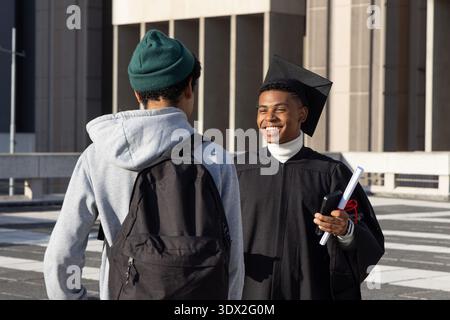 Diverse males congratulating grad in cap and gown holding diploma and phone on campus plaza Stock Photo